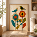 A framed vertical art print featuring a mid-century modern floral design with birds, displayed on a wooden floor in a bright, modern living room next to a window and a beige sofa.
