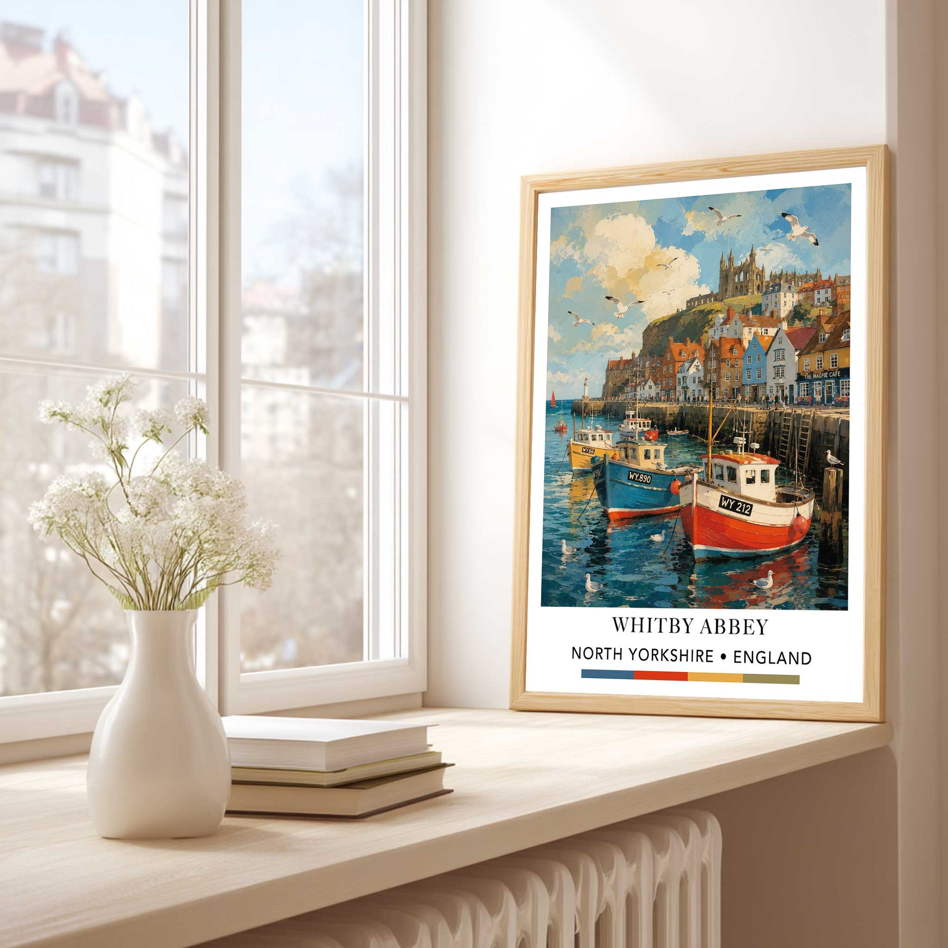 Whitby Harbour print with fishing boats, colourful waterfront buildings and Whitby Abbey in North Yorkshire  travel poster of Whitby Abbey, North Yorkshire, displayed on a windowsill next to a white vase with flowers and a stack of books. The poster features a colorful illustration of harbor boats, cliffs, and historic buildings, framed in light wood.